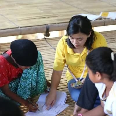 Children writing at a table in a bamboo hut while a young woman helps them