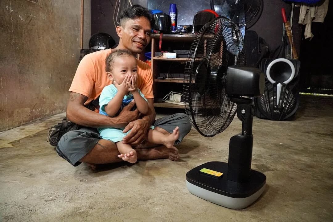 A man holding a child, sitting beside a standing solar fan