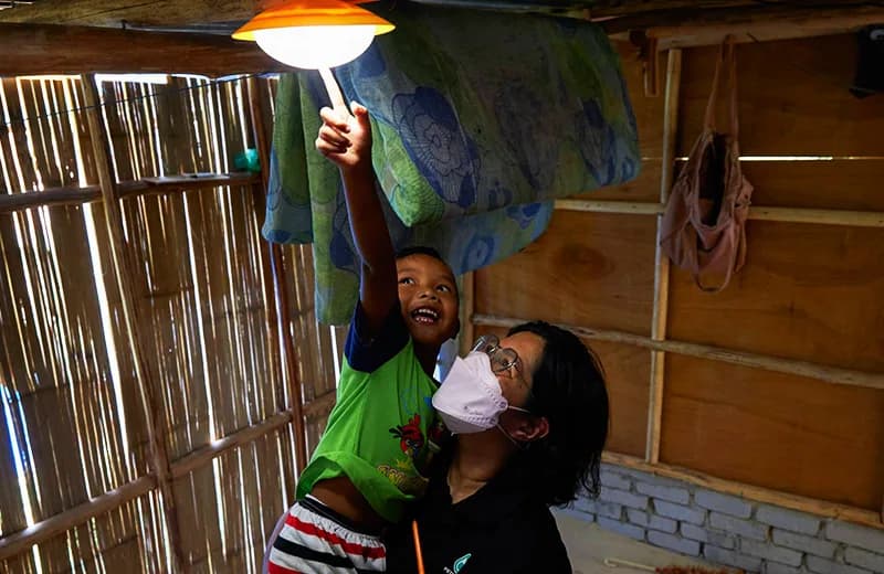 Two people inside a bamboo-walled home reaching up to a hanging solar light