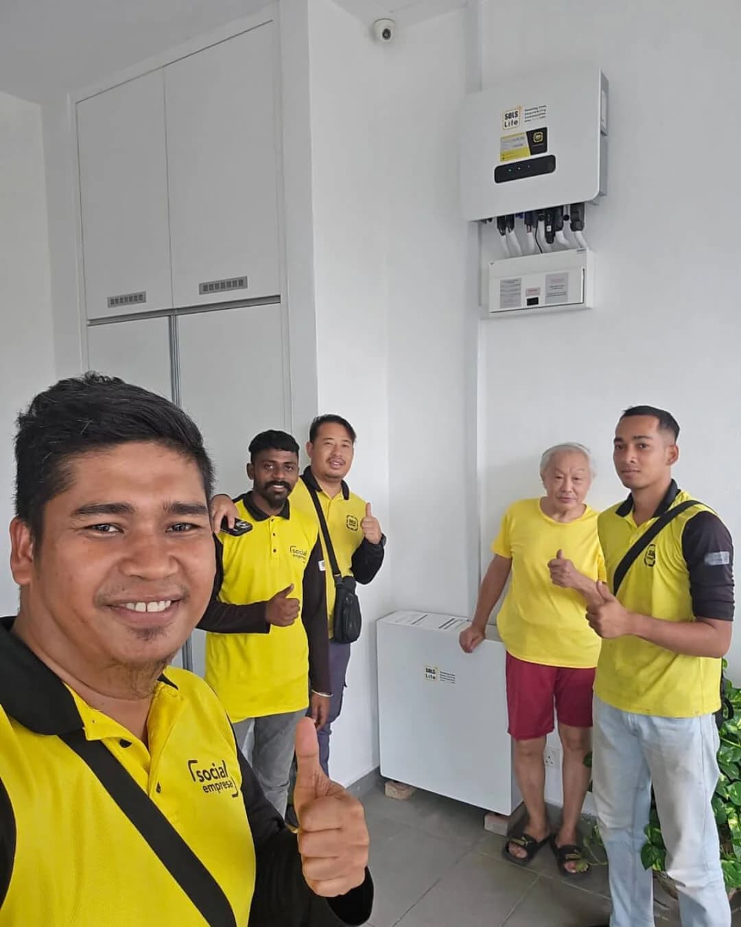 A group of people in SOLS yellow shirts beside a wall-mounted inverter and battery