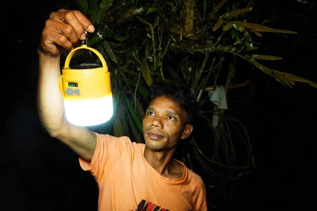 A young man holding up a lit solar lantern outdoors at night