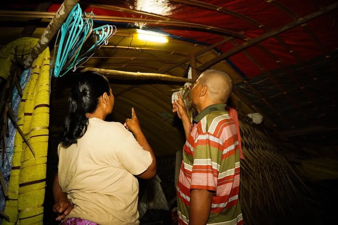 Two people inside a thatched hut lit by a hanging light