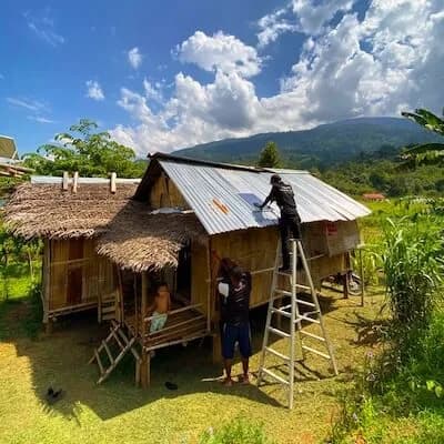 A worker on a ladder fitting a solar panel to the roof of a wooden stilt house